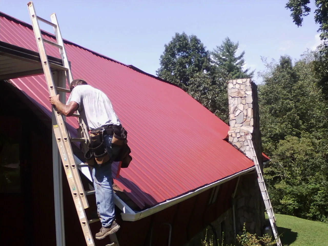 Roofer on a ladder beside a red metal roof and stone chimney — Soffit & Fascia Repair by Roofing Brothers in Brookville